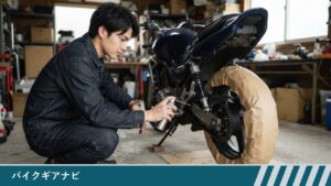 作業場で、男性がしゃがんで、タイヤを紙でおおった後輪のチェーンにスプレー缶のノズルを向けている様子
