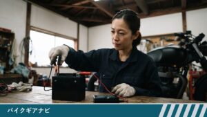 作業台のバイク用バッテリーの右の「+」端子に赤い線の赤いクリップを付け、左の「−」端子に黒い線の黒いクリップを手袋の手でつなごうとする、作業服の女性