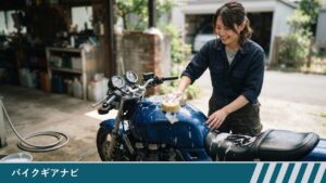 自宅の庭先で、女性が青いバイクのタンクの上面に泡をつけたスポンジを当てて洗っている様子