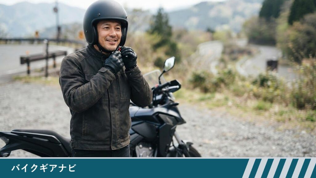 停車したバイクの横で、男性がグローブをはめた手でヘルメットのあごひもを締めている様子