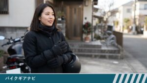 玄関先で、ネックウォーマーと黒い冬用グローブを着けた女性が、ヘルメットを抱えて手を胸の前に合わせて立っている様子