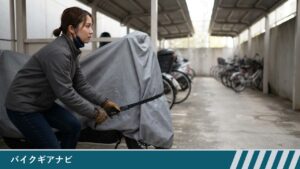 屋根付きの駐輪場で、女性がグレーのカバーをかけたスクーターの前側で、黒いベルトのバックルを引いて締めている様子