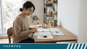 自宅の机で、女性が書類に手を置いて見ており、机の上にはほかの書類と冊子、車のキーが置いてある様子