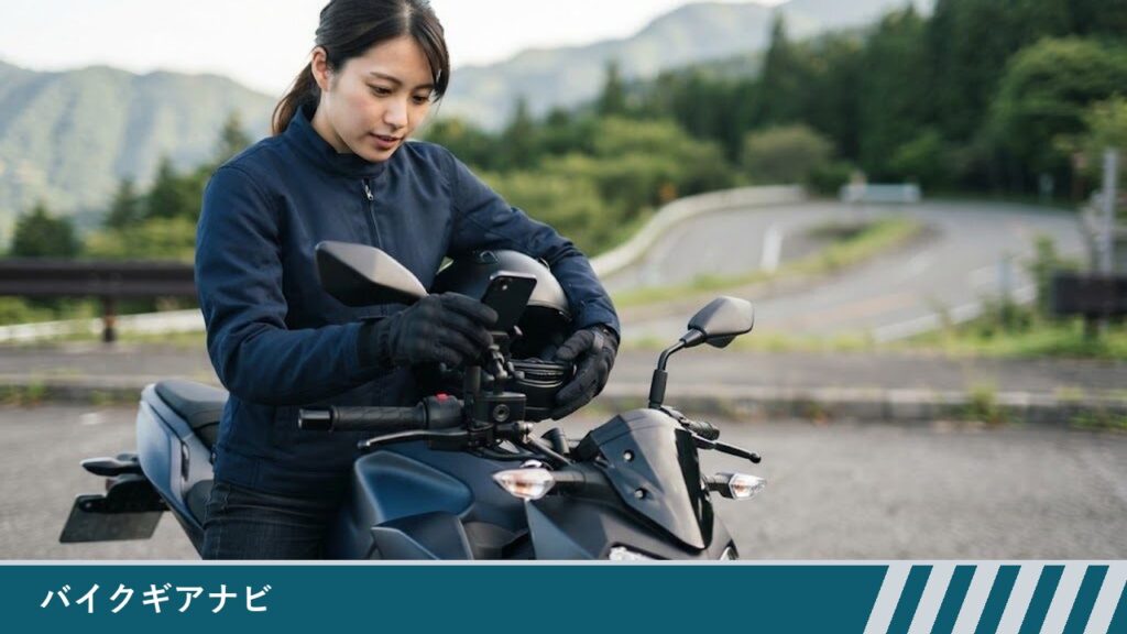 停車したバイクにまたがった若い女性ライダーが、ハンドルに付けたスマホホルダーのスマホの向きを手袋をした手で整えている様子
