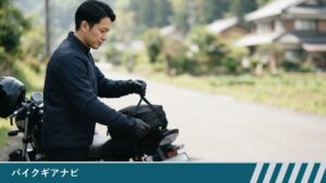 停車したバイクの後ろ席に積んだシートバッグのベルトを、男性ライダーが手袋をした手で引いて締めている様子